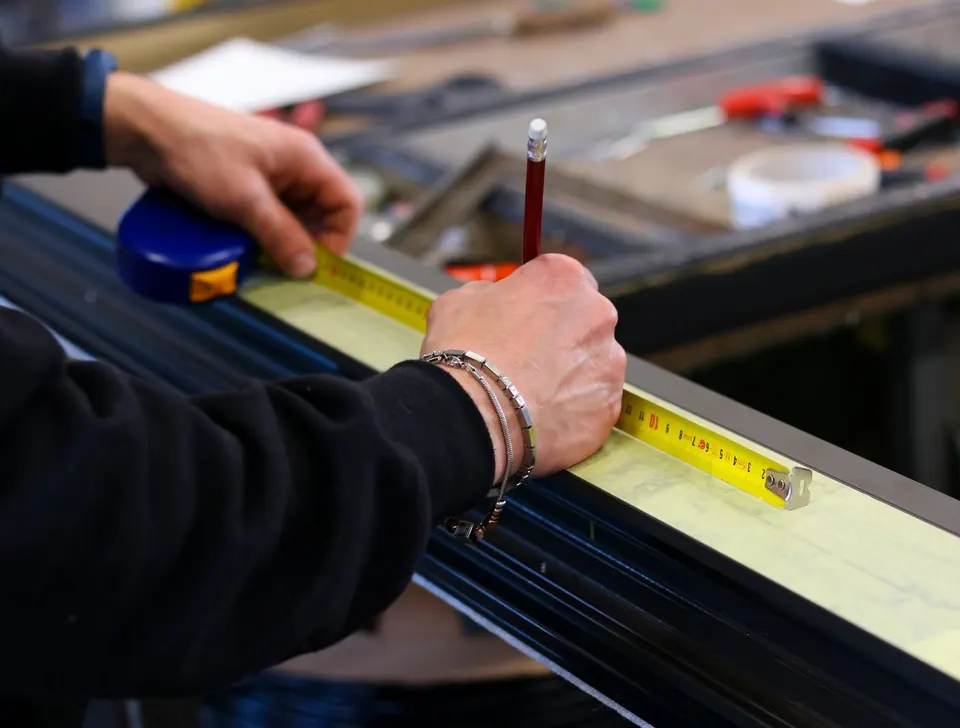 Manos midiendo con flexómetro y marcando con lápiz un perfil de aluminio en el banco del taller